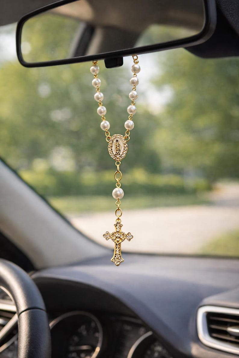 May include: A car rosary with faux pearl beads and gold-tone accents. The rosary features a cross pendant with clear crystal-like embellishments and a central medallion with a religious figure.