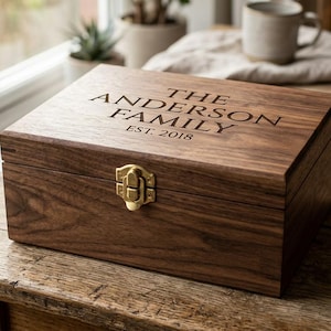 May include: A rectangular dark brown wooden keepsake box with a hinged lid and a gold-toned metal latch. The lid is engraved with the text THE ANDERSON FAMILY and EST. 2018. The box features a visible wood grain pattern and a smooth finish.