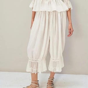 May include: Off-white linen ensemble featuring a ruffled top and cropped pants with lace trim at the ankles. The outfit is paired with beige sandals. The clothing has a vintage, romantic aesthetic, suitable for warm weather.
