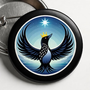Op de afbeelding: Een zwarte button met een gestileerde vogel met uitgespreide vleugels, met een gouden kroon. De vogel staat tegen een blauw-witte achtergrond met een ster. De button heeft een metalen sluiting aan de achterkant.