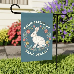 Op de afbeelding: Een tealkleurige tuinvaandel met een illustratie van een wit konijn omringd door een bloemenkrans. De vlag toont de tekst "RADICALIZED BY BASIC DECENCY" in witte letters. De vlag hangt aan een zwarte metalen standaard.