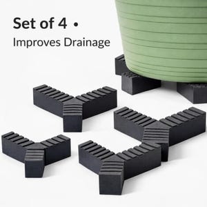 Puede incluir: Juego de cuatro elevadores negros para macetas. Cada elevador tiene forma de Y con superficies ranuradas, diseñadas para mejorar el drenaje. Una maceta verde descansa sobre los elevadores.