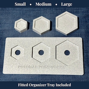Può includere: Un set di tre contenitori esagonali di dimensioni piccole, medie e grandi, insieme a un vassoio organizer abbinato. I contenitori e il vassoio sono grigio chiaro con una trama maculata. Il vassoio ha ritagli per i contenitori e il testo "POTOMAC PRINTWORKS".