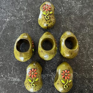 May include: Six olive green ceramic napkin rings. Each ring features a hand-painted red flower with green leaves and a yellow stem. The napkin rings have a rustic, handcrafted appearance, perfect for a nature-themed table setting.