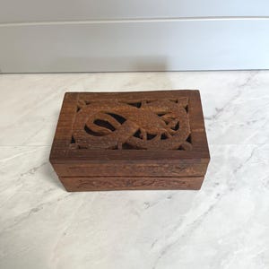 May include: A rectangular brown wooden box featuring an intricate openwork carving of a dragon on the lid. The sides of the box are also decorated with carved patterns. The wood has a natural grain and a medium brown finish throughout the piece.