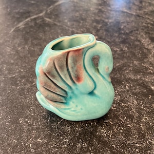 Op de afbeelding: Een kleine, turquoise keramische zwaanfiguur met een roodbruin vleugelontwerp. De zwaan heeft een gebogen nek en een kleine opening aan de bovenkant. De figuur staat op een donker, gespikkeld oppervlak.
