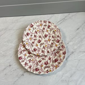 May include: Two round ceramic plates with scalloped edges, featuring a repeating pattern of dark red roses and small yellow flowers on a cream background. The plates are stacked, with the smaller plate resting on top of the larger plate.