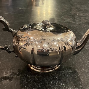 Könnte beinhalten: Eine silberne Teekanne mit kunstvollen floralen Gravuren und einem gebogenen Griff. Die Teekanne hat einen runden Körper, einen Ausguss und einen Deckel mit dekorativem Knauf. Die Teekanne steht auf einer dunklen Oberfläche.