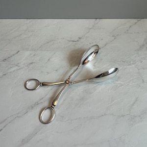 May include: Silver serving tongs with spoon-shaped ends and ring handles. The tongs are open, showing their use. The design is simple and elegant, suitable for serving various foods.