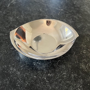 May include: A polished, silver serving bowl with an unusual, eye-shaped form. The bowl has a smooth, reflective surface and gently curved sides, with small handles at each end. The bowl is likely made of metal and is suitable for serving food.