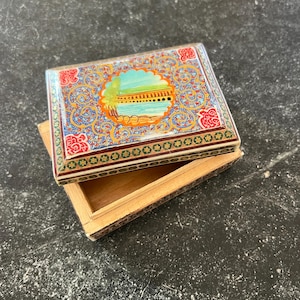 May include: A rectangular, open wooden box with a decorative lid. The lid features a colorful, ornate design with a central landscape scene in a circular frame. The box is partially open, revealing the plain wooden interior.