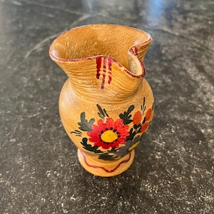 Könnte beinhalten: Kleine, hellbraune Keramikvase mit ausgestelltem Rand und rundem Körper. Die Vase ist mit handgemalten Blumendesigns in Rot, Gelb und Schwarz verziert. Der Sockel hat eine dekorative rot-braune Verzierung.