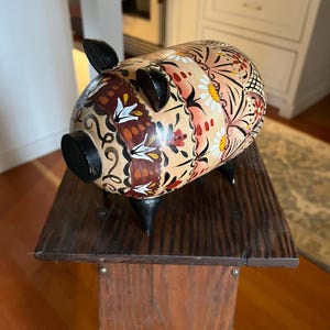 May include: A decorative piggy bank with a floral design in shades of peach, red, and white. The pig has black ears, snout, and legs. It is displayed on a dark wooden stand.