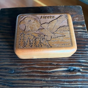 May include: A wooden box with a carved design featuring an eagle, trees, and the word "Alaska." The eagle is depicted in flight, carrying a fish. The box is rectangular with a natural wood finish.