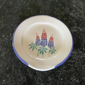 May include: A white ceramic spoon rest with a blue rim and a floral design. The design features three stylized purple and red flowers with green leaves. The spoon rest has a curved indentation for holding utensils.