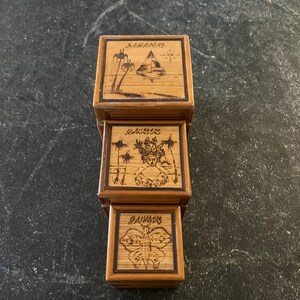 May include: Stacked wooden boxes with etched designs and the word "Bahamas". The top box features palm trees and a pyramid. The middle box shows a person with a headdress. The bottom box has a butterfly.