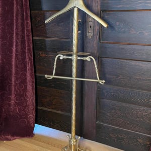 May include: A vintage brass valet stand with a coat hanger, tie rack, and a shoe rest. The stand has a decorative ball finial and a detailed base. The stand is approximately 65 inches tall.