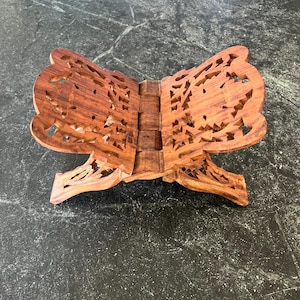 May include: A wooden book stand with intricate, openwork designs. The stand is made of a warm, reddish-brown wood and features a hinged design for easy folding and storage. The detailed carvings add a decorative element to this functional item.