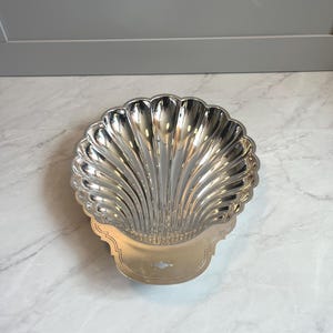 May include: A silver-colored, shell-shaped dish with a scalloped rim and a decorative, elongated handle. The dish has a reflective surface and is likely made of metal. The design is reminiscent of Art Deco style.