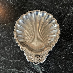 May include: A silver-colored, shell-shaped dish with a scalloped edge and ornate floral detailing. The dish has a reflective surface and is likely made of metal. The design is reminiscent of antique serving pieces.