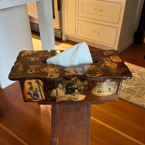 May include: A decorative tissue box with a dark brown, glossy finish. The box is adorned with vintage black and white photographs and sits atop a wooden stand. White tissues are visible at the top. The box is a unique home decor item.