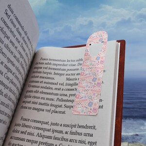 May include: A pink bookmark with a heart-shaped hole is placed in a book. The bookmark has a repeating pattern of glasses, books, magnifying glasses, and animal faces. The book is open, with text visible on the pages.