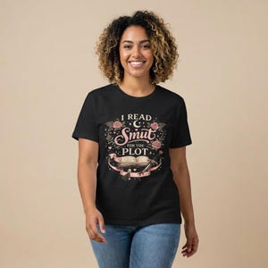May include: Black t-shirt with the text "I READ SMUT for the PLOT" in a decorative design featuring roses, a crescent moon, and an open book. The design is in pink, black, and white.