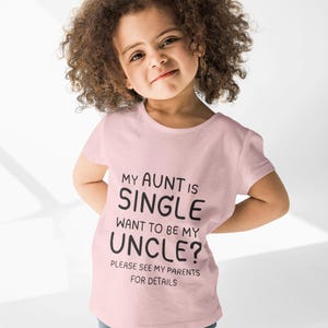 May include: A light pink t-shirt with the text "MY AUNT IS SINGLE WANT TO BE MY UNCLE? PLEASE SEE MY PARENTS FOR DETAILS" in black. The shirt is worn by a young child with curly brown hair and blue jeans.