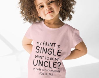 May include: A light pink t-shirt with the text "MY AUNT IS SINGLE WANT TO BE MY UNCLE? PLEASE SEE MY PARENTS FOR DETAILS" in black. The shirt is worn by a young child with curly brown hair and blue jeans.
