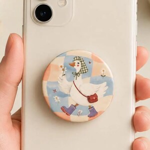 May include: A white smartphone featuring a circular phone grip on the back. The grip displays an illustration of a white goose wearing a green checkered bonnet, purple boots, and a red crossbody bag, set against a light blue and peach checkered background.