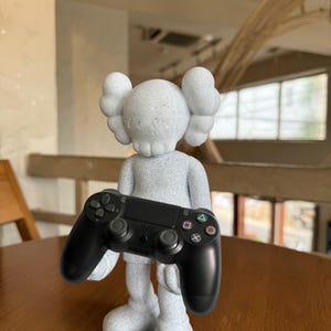 May include: A gray sculpture of a figure holding a black PlayStation controller. The figure has a skull-like face and large, rounded ears. The controller has the standard button layout with a directional pad, face buttons, and analog sticks.