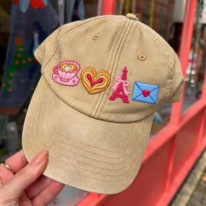 Puede incluir: Una gorra de béisbol de color tostado con cuatro parches bordados: una taza de café rosa con arte latte, un pastel en forma de corazón, una Torre Eiffel rosa con un lazo y un sobre azul claro con un corazón rojo.