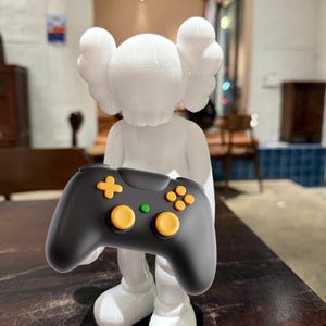 May include: A white figurine of a cartoon character holding a black video game controller. The controller has orange and green buttons. The figurine has large, round ears and is standing on a wooden surface.