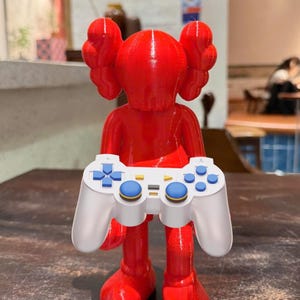 May include: A vibrant red figurine holding a white and blue video game controller. The figurine has a unique design with large, rounded ears and a smooth, glossy finish. The controller features blue and white buttons.