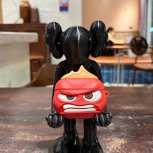 May include: A black figurine holding a red and white AirPod case. The AirPod case is designed to look like the character Anger from the movie Inside Out. The figurine has large ears and a stylized appearance.