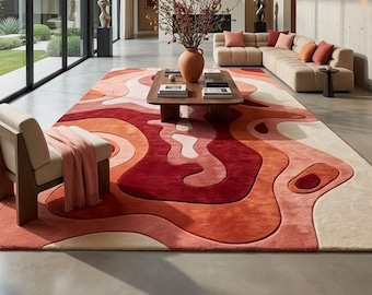 May include: A large, abstract area rug with a layered, wavy design in shades of red, orange, and cream. The rug is the focal point of a modern living room, complemented by a beige sectional sofa, wooden coffee table, and a neutral-toned armchair.