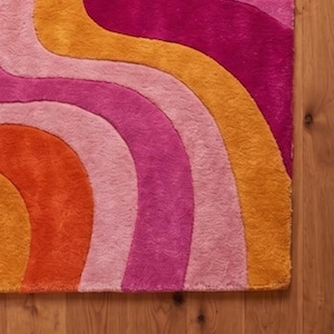 May include: A rectangular area rug featuring a series of wavy, curved stripes in vibrant shades of orange, light pink, magenta, and deep pink. The rug has a thick, plush texture and is arranged in a repeating pattern of flowing, organic lines.