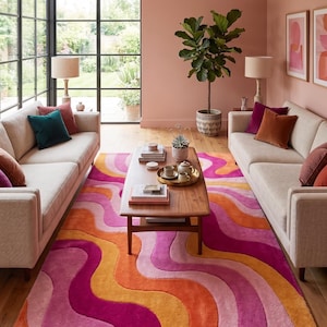 May include: A rectangular area rug featuring a wavy, multi-colored pattern in shades of bright pink, magenta, orange, and light peach. The rug is positioned on a wooden floor between two beige sofas, with a wooden coffee table placed on top.
