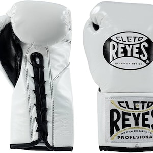Puede incluir: Guantes de boxeo de cuero blanco con detalles y cordones negros. Los guantes presentan el logo "CLET0 REYES" en negro y dorado. También se ven las palabras "HECHO EN MEXICO" y "PROFESIONAL".