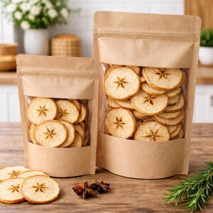 Może przedstawiać: Dwa stojące woreczki z papieru kraft z przezroczystymi okienkami wypełnione suszonymi plasterkami jabłka. Plasterki jabłka są jasno brązowe z gwieździstym rdzeniem. Kilka plasterków leży na drewnianej powierzchni z anyżem gwiazdkowym i rozmarynem.