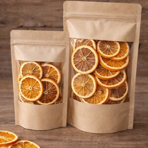 May include: Two kraft paper bags with clear windows, filled with dried orange slices. The oranges are a mix of light and dark orange, with visible textures. The bags are on a wooden surface, with a few orange slices scattered around.