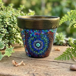 May include: A round terracotta plant pot featuring a hand-painted mandala design with intricate multicolored dots in shades of blue, green, purple, and orange. The rim of the pot is painted in a solid metallic gold finish, contrasting with the dark base.