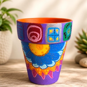 May include: A hand-painted terracotta plant pot featuring a vibrant purple rim decorated with colorful geometric shapes and a main body displaying a large, intricate blue and yellow floral mandala pattern with orange and pink accents.