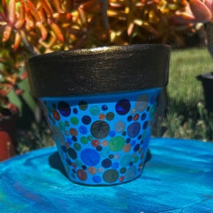 May include: A hand-painted ceramic flower pot with a blue base and a black rim. The pot is decorated with various sized colorful polka dots in shades of blue, green, orange, and silver. The pot is sitting on a blue surface.