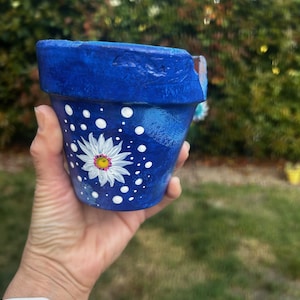 May include: A hand-painted blue ceramic flower pot with a white daisy design and white polka dots. The daisy has a yellow center. The pot is held against a blurred green background.