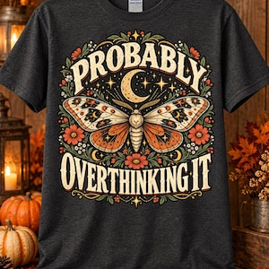 Peut inclure: Un t-shirt gris anthracite à manches courtes avec une impression graphique représentant un papillon de nuit aux ailes orange et crème, entouré de fleurs, d'étoiles et d'un croissant de lune. Le texte PROBABLY OVERTHINKING IT est imprimé en crème.