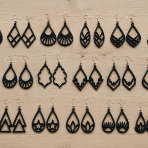 Op de afbeelding: Een collectie van achtentwintig paar zwarte oorbellen gerangschikt in vier rijen. Elk paar heeft een uniek geometrisch, bloemvormig of abstract uitgesneden ontwerp. De oorbellen hangen aan zilverkleurige metalen haakjes en tonen diverse vormen.