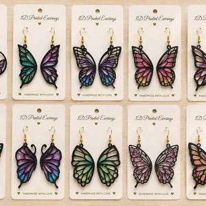 Peut inclure: Dix paires de boucles d'oreilles imprimées en 3D présentées sur des cartes blanches marquées 3D Printed Earrings et Handmade with Love. Chaque paire présente des cadres noirs avec des motifs d'ailes de papillon irisés et des crochets dorés.