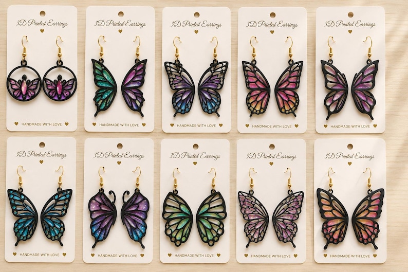 以下が含まれることがあります： 3D Printed EarringsおよびHandmade with Loveと記された白い台紙に展示された10組の3Dプリントイヤリング。各ペアは、黒いフレームにカラフルで虹色に輝く蝶の羽のデザインが施され、ゴールドトーンの金属製イヤリングフックが付いています。