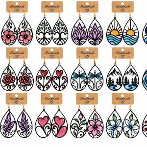 Op de afbeelding: Een collectie van achttien paar druppelvormige oorbellen met zwarte omlijningen en diverse kleurrijke ontwerpen zoals bloemen, bomen, harten, landschappen en hemelse motieven, elk paar bevestigd op een bruine kartonnen displaykaart.